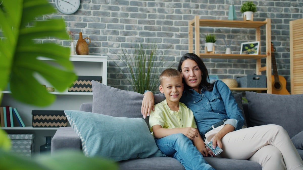 Mom and kid adorable blond boy are watching TV together laughing talking bonding in modern apartment together. People, technology and lifestyle concept.